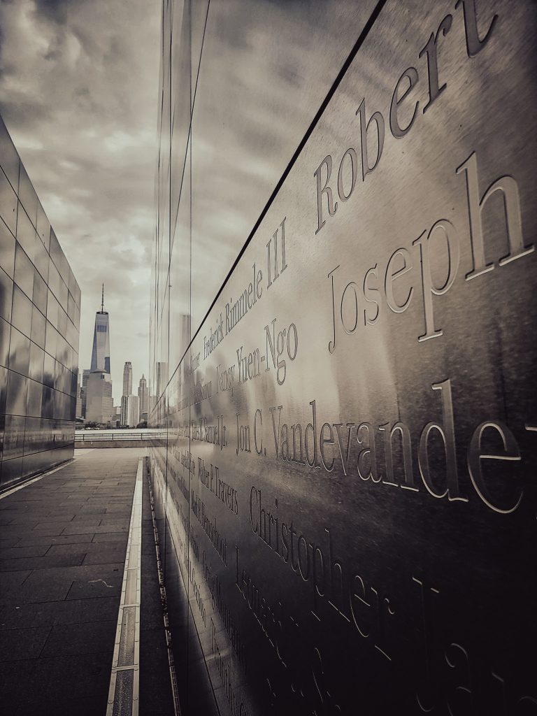 Reflective memorial with inscribed names, One World Trade Center skyline view.