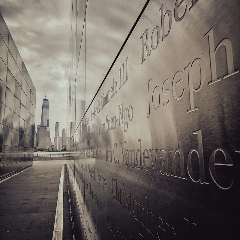 Reflective memorial with inscribed names, One World Trade Center skyline view.